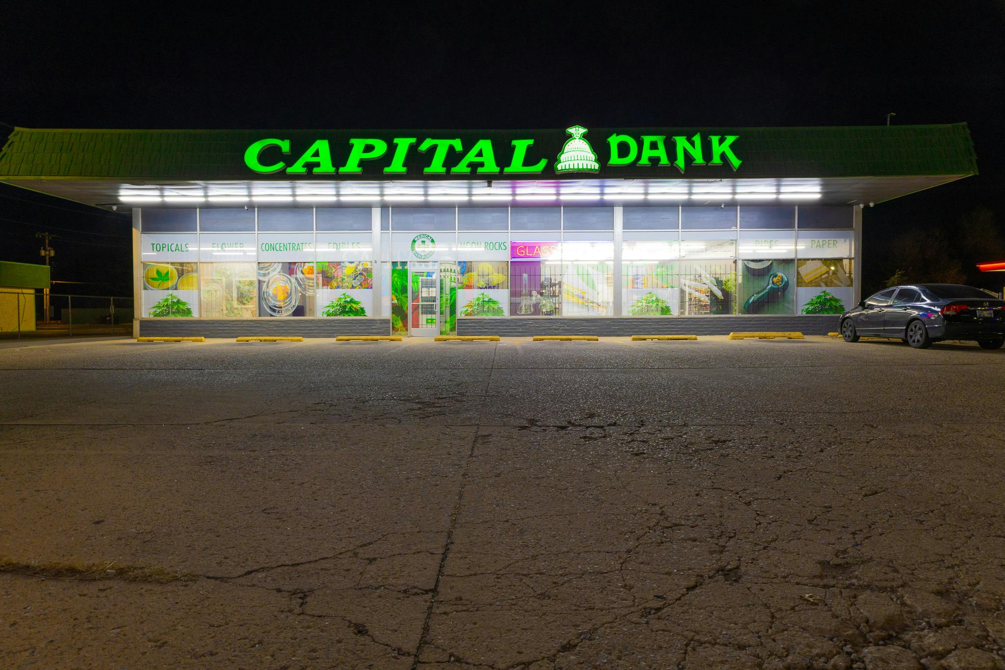 Capital Dank dispensary storefront in Midwest City, Oklahoma, lit up at night with bright green signage and product categories visible in the windows.